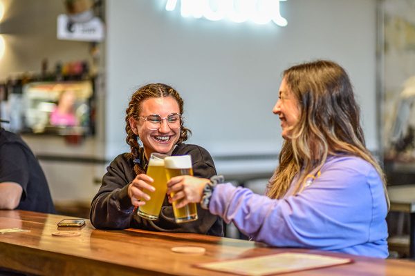 Two smiling women clinking glasses of beer at a bar.